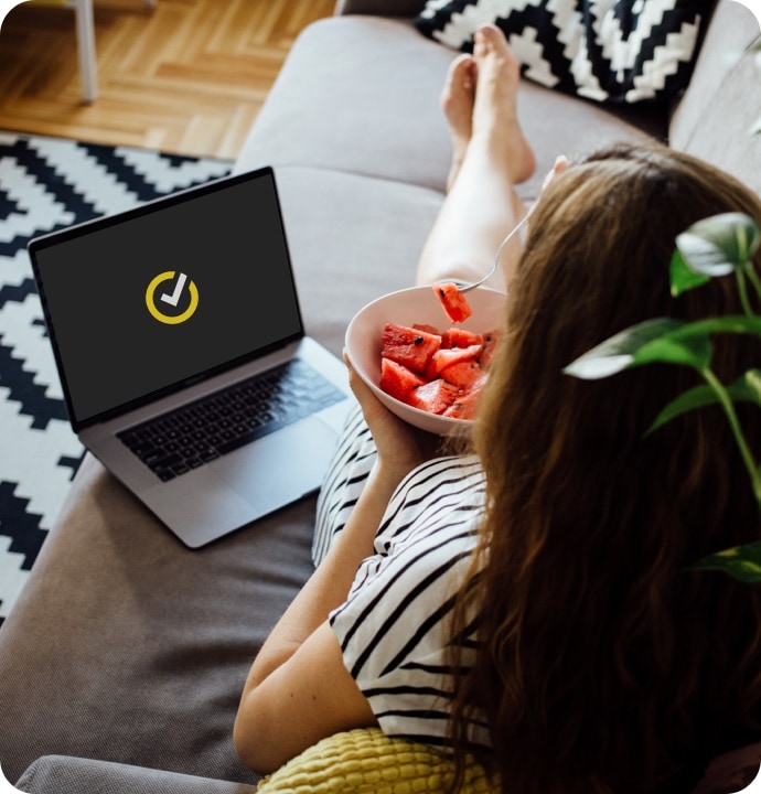 Kanapén fekvő nő, aki gyümölcsöt eszik egy tálból, mellette egy laptop, rajta a Norton logójával