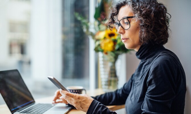 Egy nő egy mobiltelefonra néz egy laptop előtt